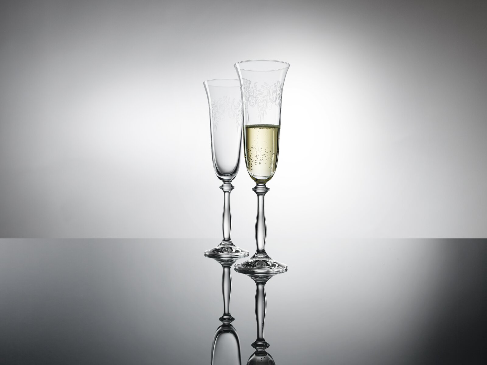 Zwei elegante Sektkelche mit feinem Muster, einer gefüllt mit Sekt, auf spiegelnder Oberfläche vor grauem Hintergrund, seitliche Perspektive.