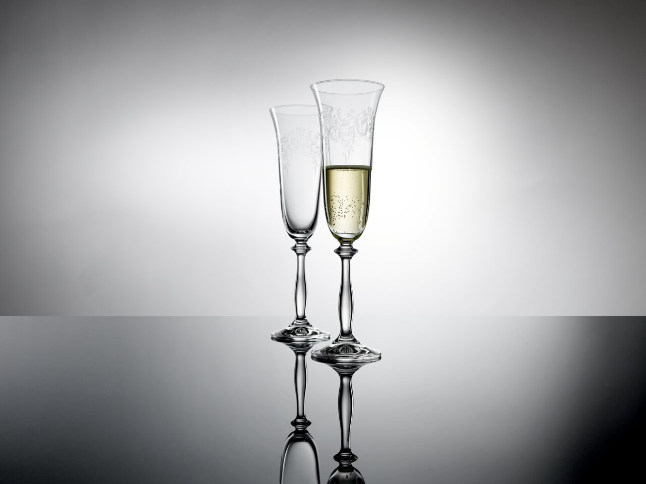 Zwei elegante Sektkelche mit feinem Muster, einer gefüllt mit Sekt, auf spiegelnder Oberfläche vor grauem Hintergrund, seitliche Perspektive.
