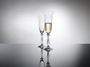 Zwei elegante Sektkelche mit feinem Muster, einer gefüllt mit Sekt, auf spiegelnder Oberfläche vor grauem Hintergrund, seitliche Perspektive.