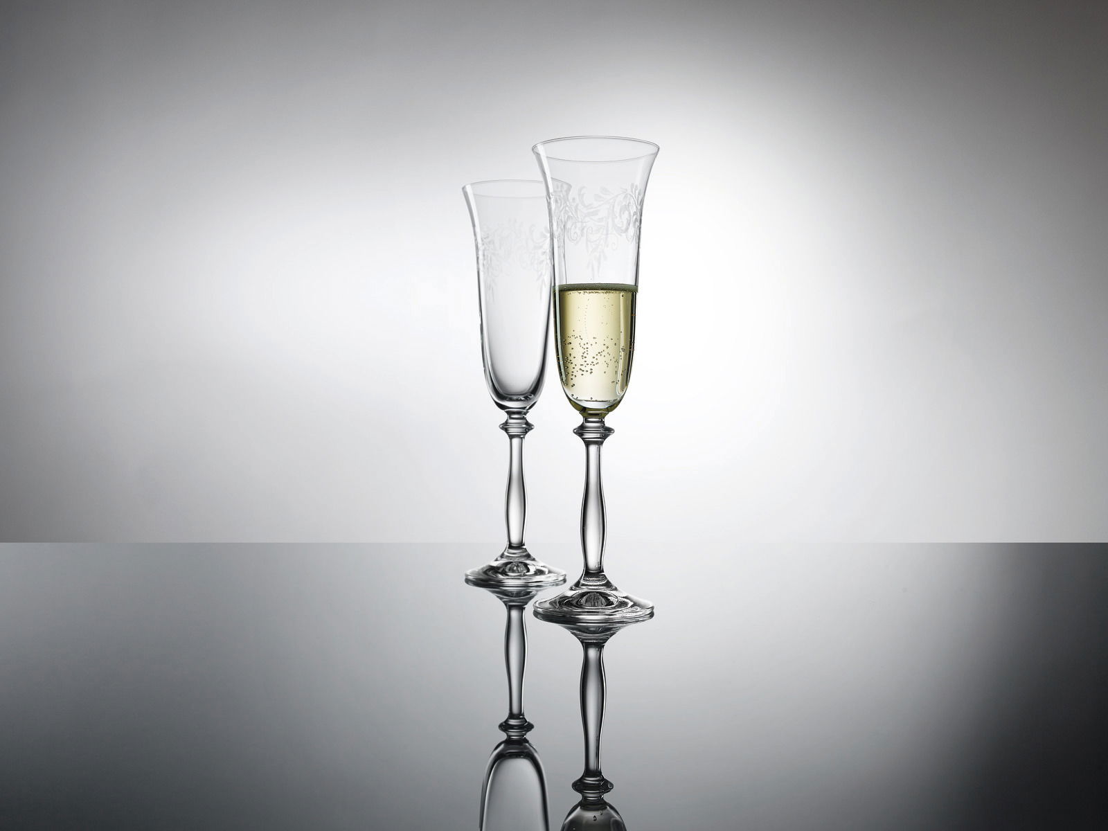 Zwei elegante Sektkelche mit feinem Muster, einer gefüllt mit Sekt, auf spiegelnder Oberfläche vor grauem Hintergrund, seitliche Perspektive.