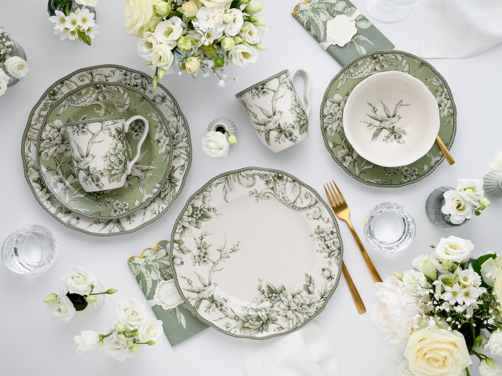 Draufsicht (Flatlay) auf ein elegantes grün-weißes Porzellan-Tafelservice mit floralen und Vogel-Motiven: Teller, Schale und Tasse, kombiniert mit goldfarbenem Besteck, Servietten und weißen Blumen auf hellem Tisch.