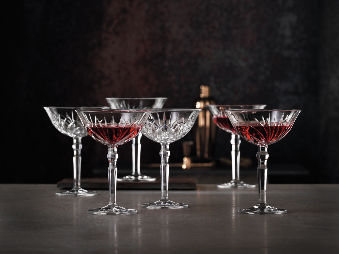 Sechs elegante Cocktailschalen aus Kristallglas mit rotem Getränk, auf einer Tischoberfläche in stilvoller Umgebung, seitliche Perspektive.