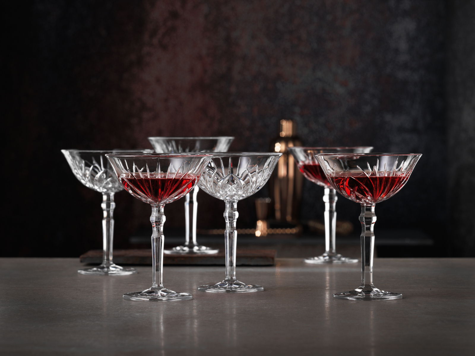 Sechs elegante Cocktailschalen aus Kristallglas mit rotem Getränk, auf einer Tischoberfläche in stilvoller Umgebung, seitliche Perspektive.