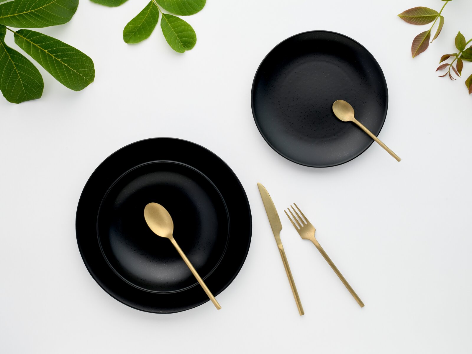 Draufsicht (Flatlay) auf schwarzes Keramikgeschirr mit zwei Tellern/Schalen und goldfarbenem Besteck auf weißem Hintergrund, dekoriert mit grünen Blättern