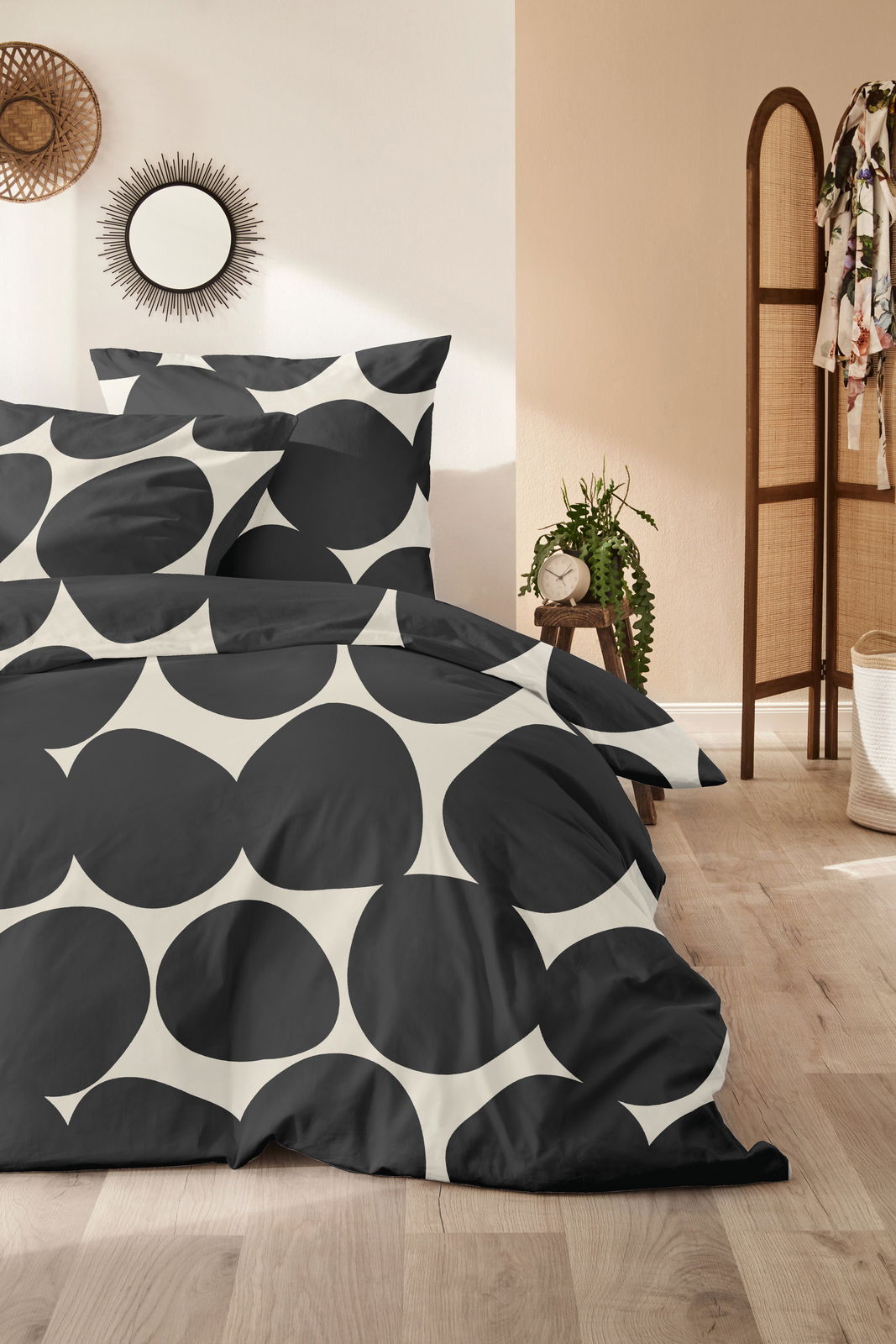 Bettwäsche-Set mit großem, schwarzem Kreismuster auf weißem Hintergrund, im Schlafzimmer mit Holzboden und Pflanzen dekoriert, aus der Perspektive von vorne.