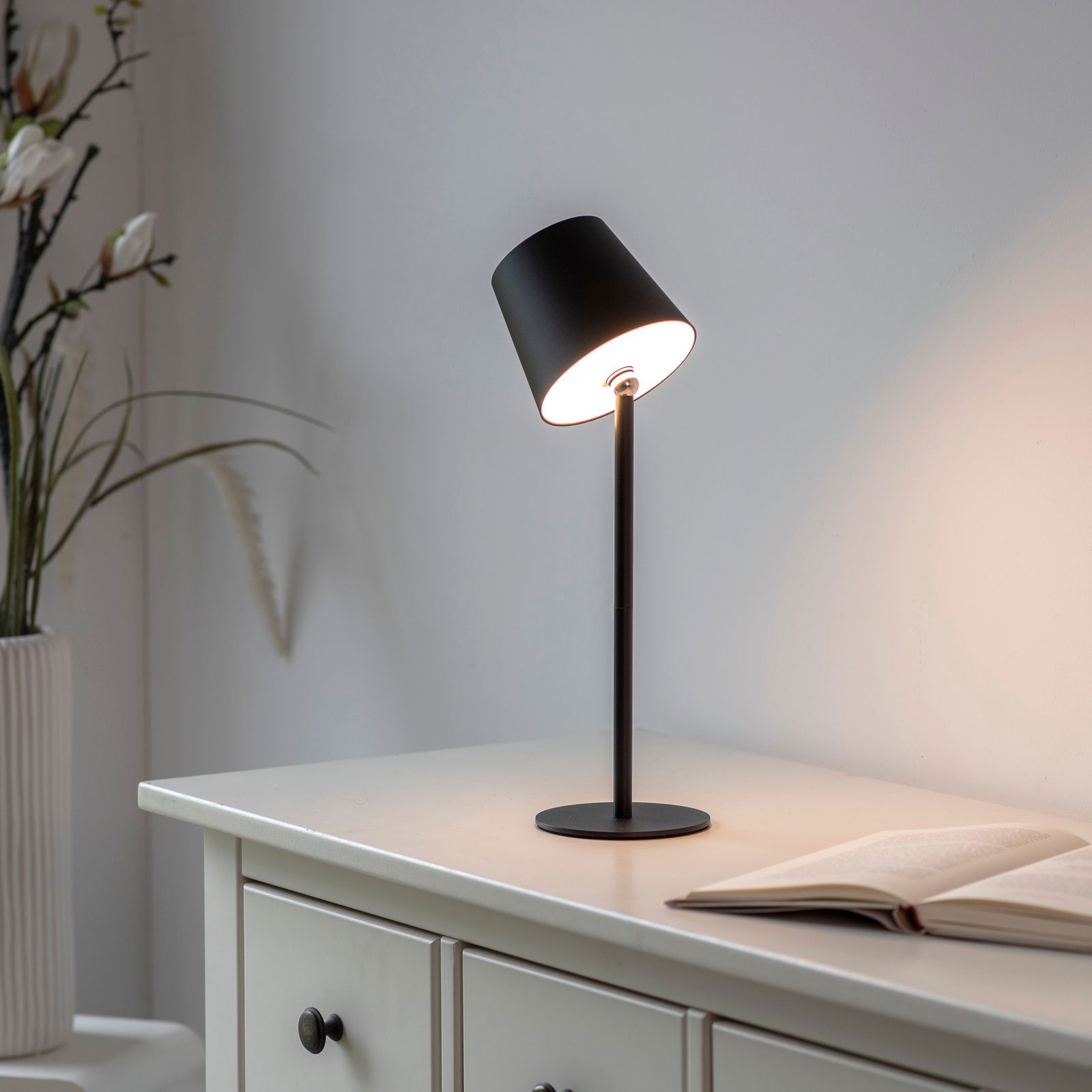 Schwarze Akku-Tischleuchte BOTERIA auf einem weißen Schrank, seitliche Perspektive, mit einem geöffneten Buch daneben.