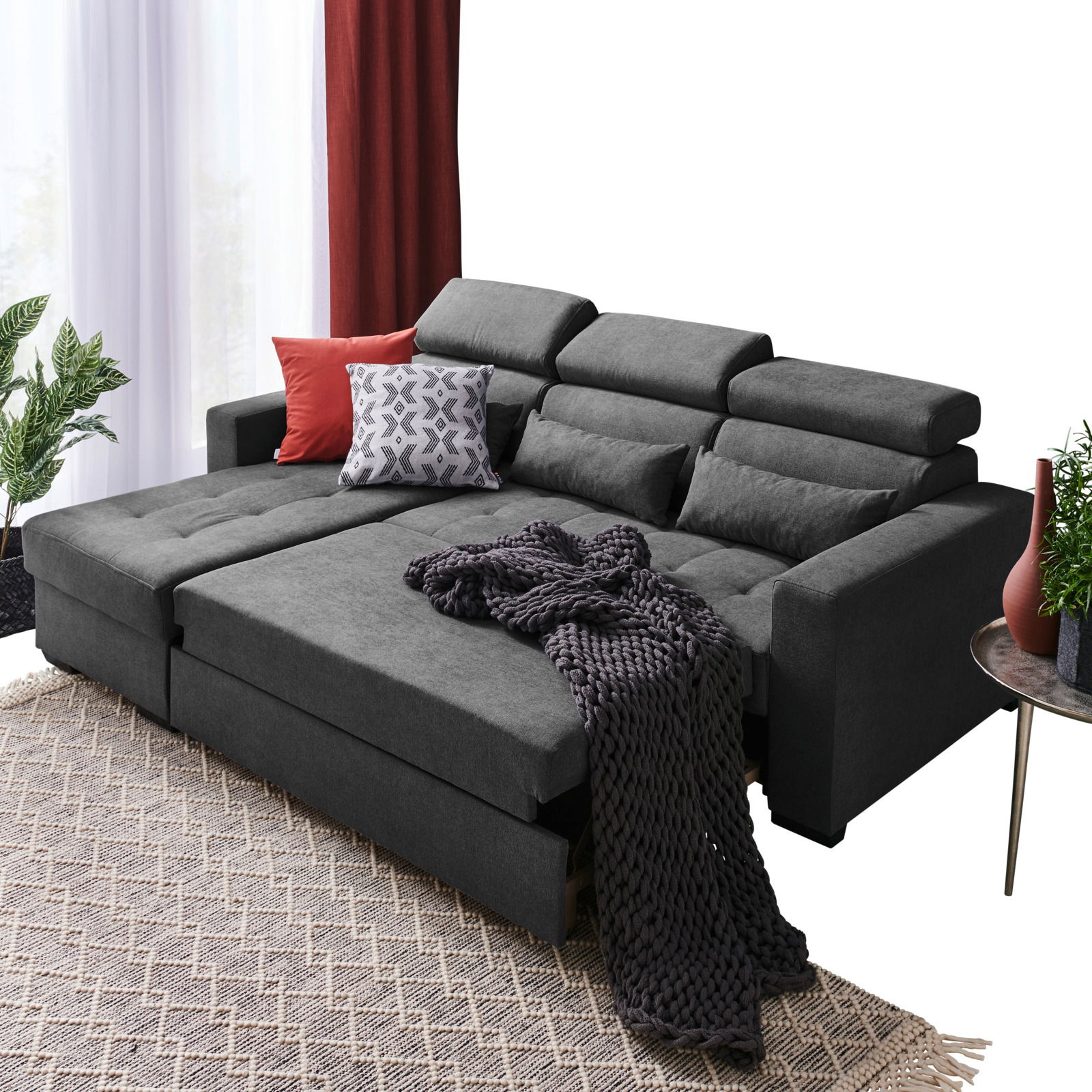 Graue Rundecke (L) mit verstellbaren Kopfstützen, dekoriert mit roten und gemusterten Kissen, aus der Perspektive von vorne links fotografiert.