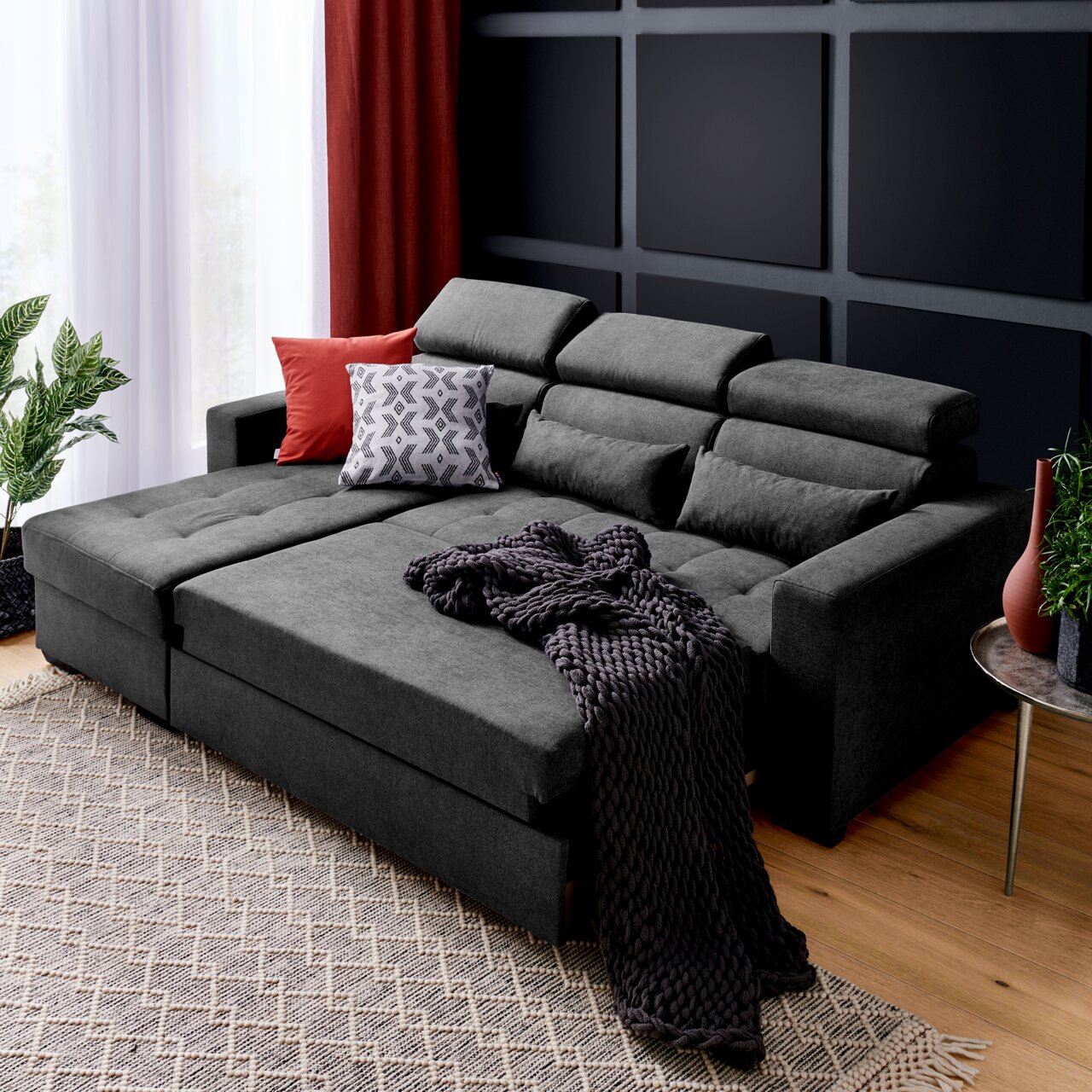 Graue Rundecke (L) mit verstellbaren Kopfstützen, dekoriert mit roten und gemusterten Kissen, aus der Perspektive von vorne links fotografiert.