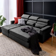 Graue Rundecke (L) mit verstellbaren Kopfstützen, dekoriert mit roten und gemusterten Kissen, aus der Perspektive von vorne links fotografiert.