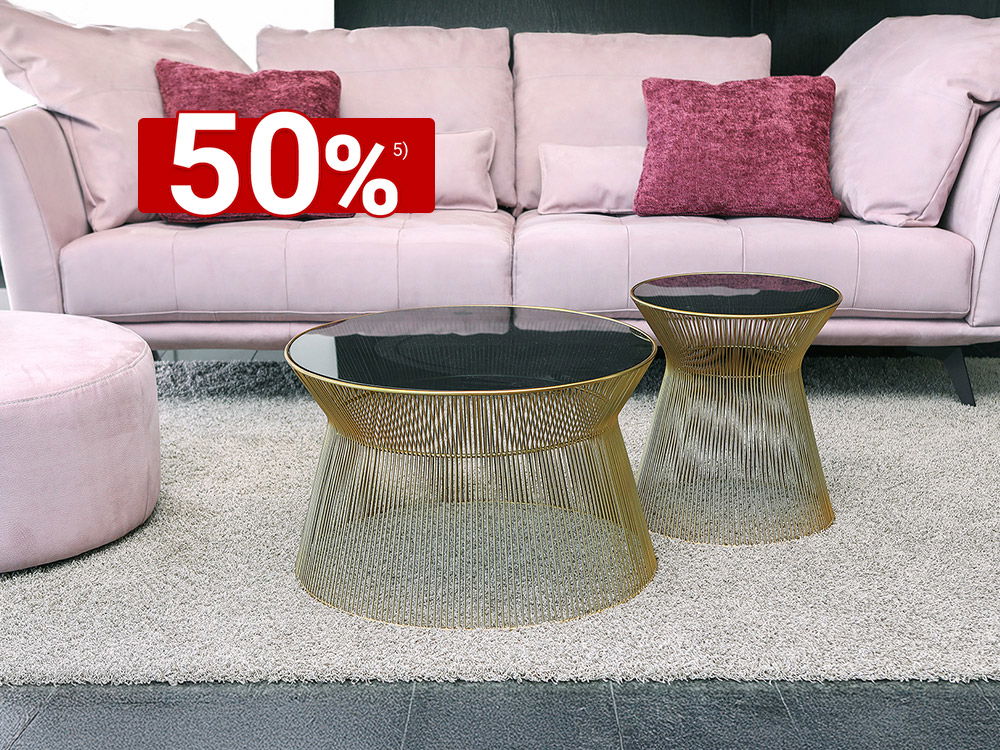 Wohnzimmer mit rosafarbenem Sofa, violetten Kissen und zwei goldenen Draht-Couchtischen mit dunkler Glasplatte auf hellem Teppich