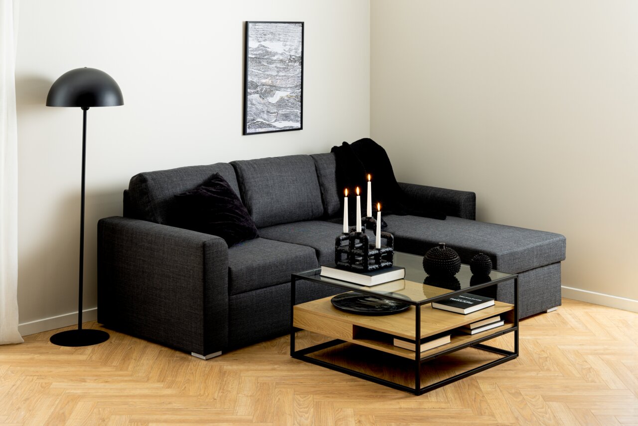 Moderner Couchtisch mit Glasplatte und Holzablage, umgeben von einem schwarzen Metallrahmen, in einem stilvollen Wohnzimmer mit grauem Sofa und Stehlampe. Perspektive von vorne.