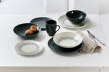 Tafelservice Cecina Graphit 18-teilig, bestehend aus Tellern, Schalen und Tassen in einem modernen Design mit grauen und weißen Akzenten, auf einem Tisch von oben fotografiert.