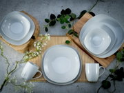 Weißes Geschirr-Set mit Tellern, Schalen und Tassen auf Holzbrett und Bambusmatte mit grünen Zweigen, Draufsicht von oben.