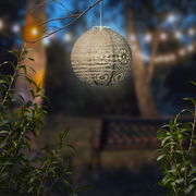 Solarbetriebene Laterne in Kugelform, die in einem Garten bei Dämmerung von einem Ast hängt, mit dekorativem Muster und warmem Licht.