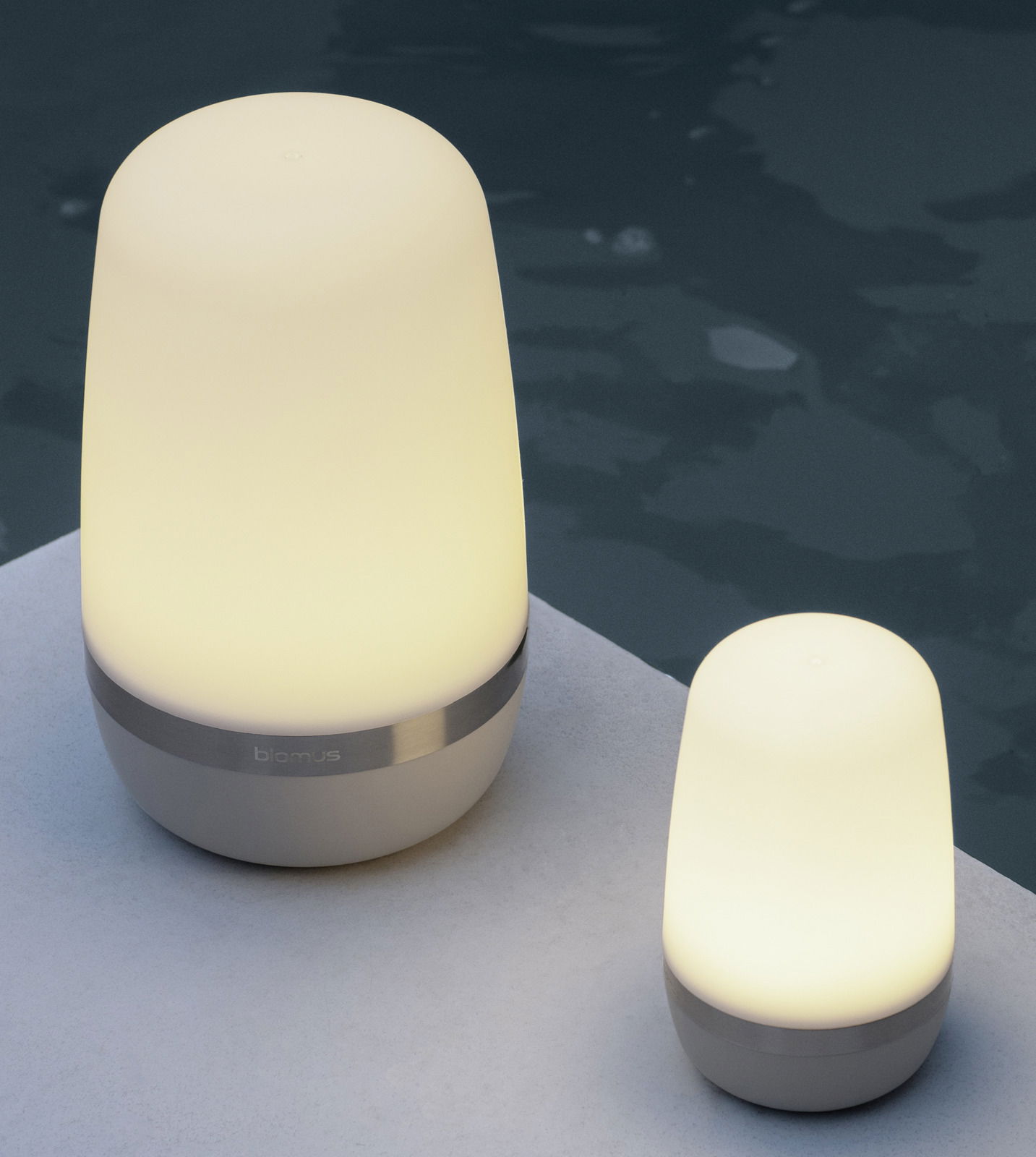 Zwei LED-Outdoorleuchten SPIRIT von Blomus, eine größere und eine kleinere, auf einer hellen Oberfläche neben einem Pool, seitliche Perspektive.