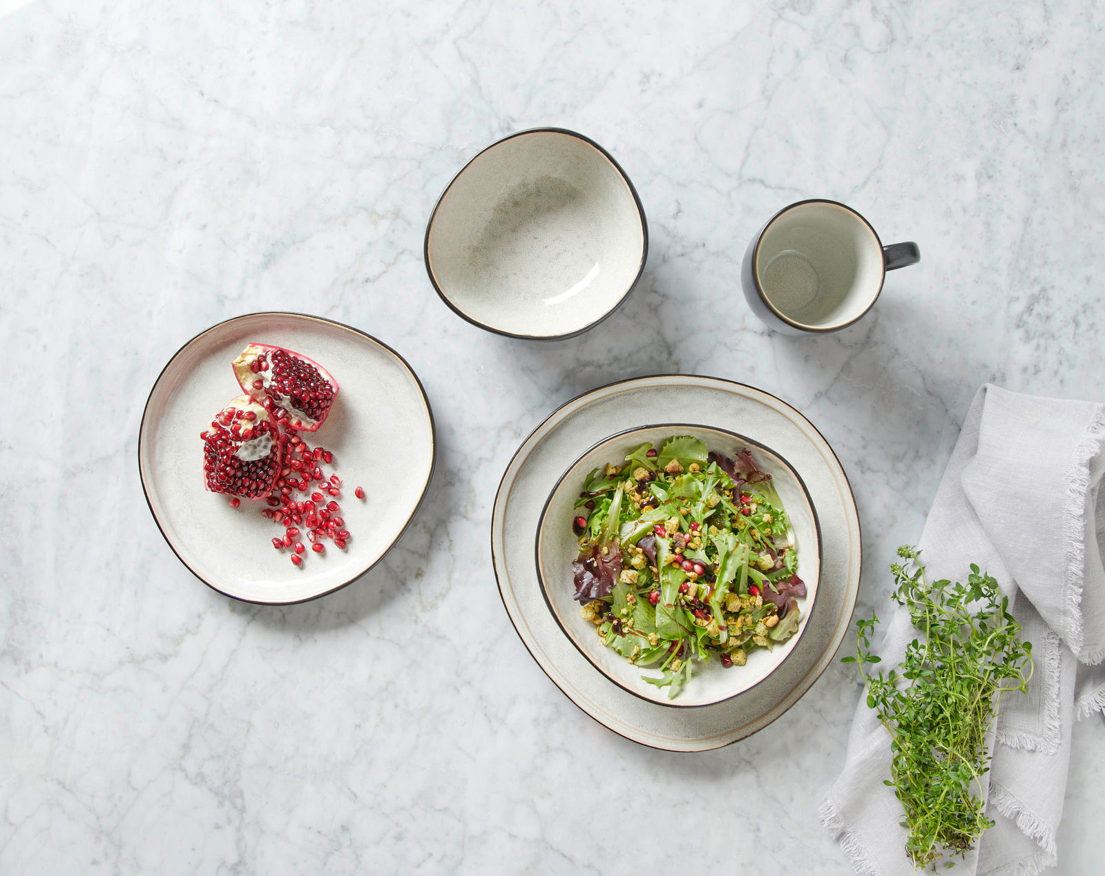Vogelperspektive auf ein graues Kombiset mit Schalen, Teller und Tasse, dekoriert mit Salat und Granatapfel auf einem Marmortisch.
