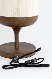 Detailaufnahme einer Tischlampe mit Holzoptik-Fuß und weißem, strukturiertem Lampenschirm; vorne liegt ein schwarzes Textilkabel mit Schalter und EU-Stecker, Perspektive: seitliche Nahaufnahme auf den unteren Lampenbereich.