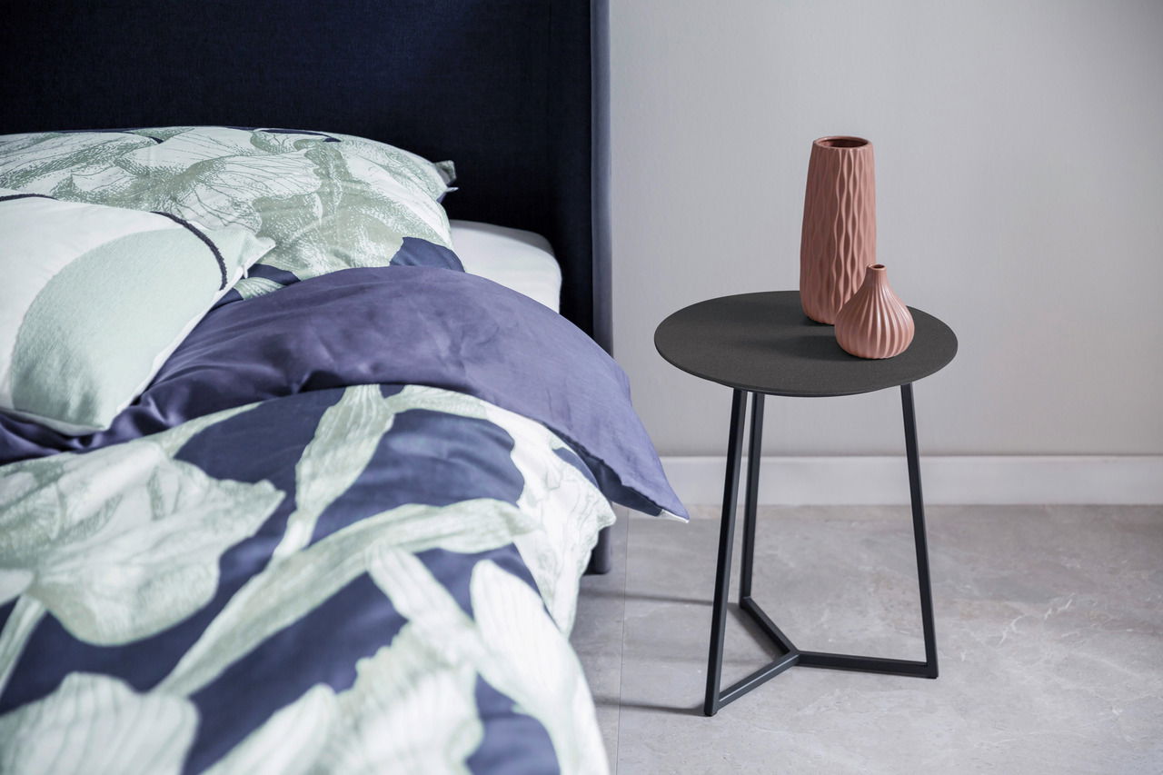 Moderner Beistelltisch mit schwarzer runder Tischplatte und geometrischem Metallgestell, dekoriert mit zwei strukturierten Vasen, seitliche Perspektive neben einem Bett.