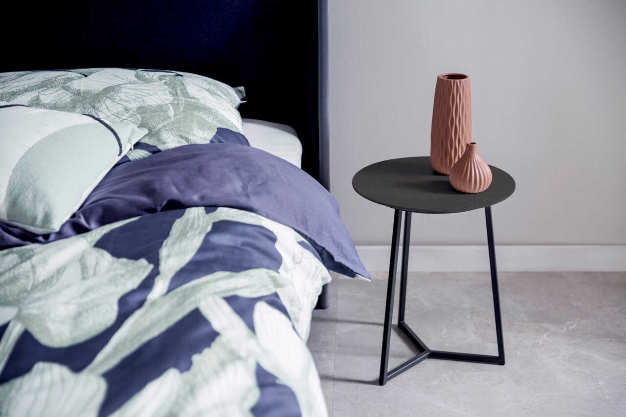 Moderne bijzettafel met een zwart, rond tafelblad en een geometrisch metalen onderstel, versierd met twee gestructureerde vazen, gezien vanuit de zijkant naast een bed.