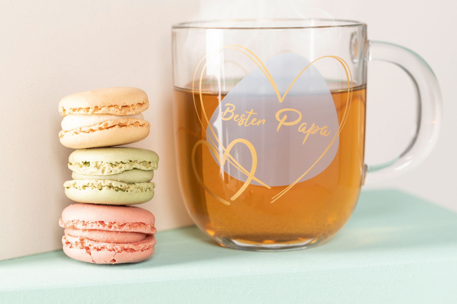 Glas-Tasse mit der Aufschrift 'Bester Papa' in Herzform, gefüllt mit Tee, neben einem Stapel bunter Macarons, seitliche Perspektive