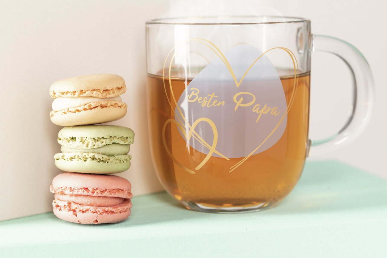 Leonardo Geschenktasse Bester Papa EMOZIONE Glas-Tasse mit der Aufschrift 'Bester Papa' in Herzform, gefüllt mit Tee, neben einem Stapel bunter Macarons, seitliche Perspektive