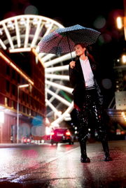 Frau mit gemustertem Regenschirm in einer nächtlichen Stadt, im Hintergrund ein beleuchtetes Riesenrad. Perspektive von vorne.