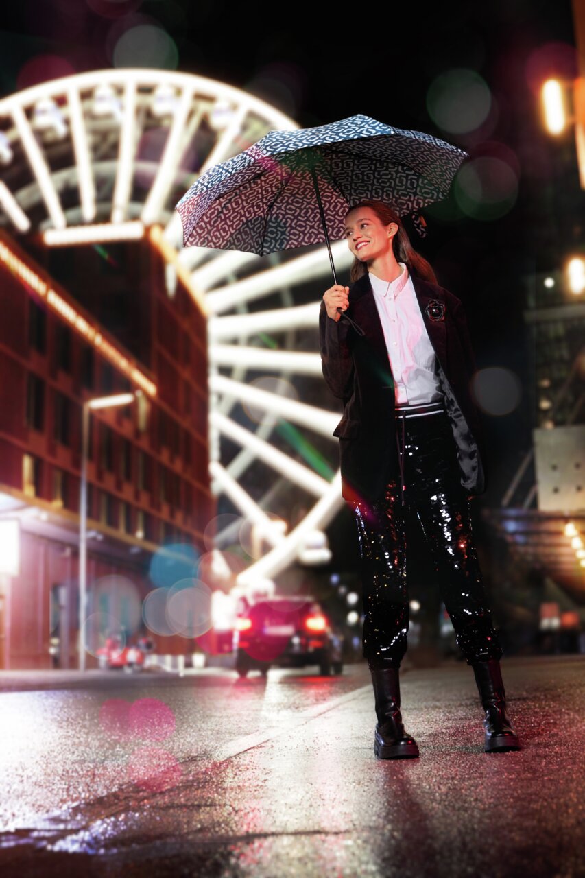 Frau mit gemustertem Regenschirm in einer nächtlichen Stadt, im Hintergrund ein beleuchtetes Riesenrad. Perspektive von vorne.