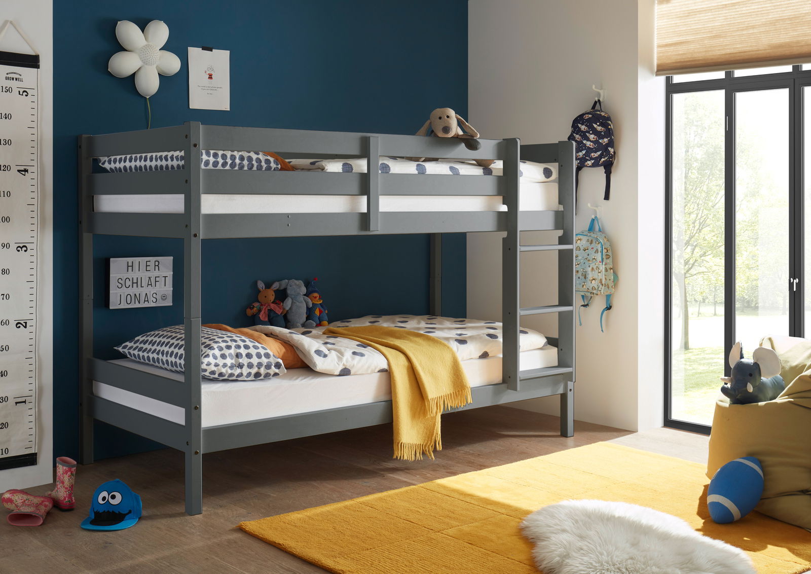 Graues Etagenbett in einem Kinderzimmer mit blauer Wand, dekoriert mit Kuscheltieren und bunten Kissen, seitliche Perspektive.