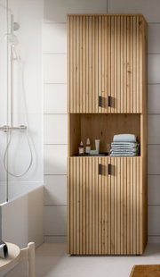 Hoher Badezimmerschrank aus Holz mit vertikaler Lamellenfront, zwei Türen oben und unten sowie offenem Mittelregal mit Handtüchern und Pflegeprodukten, frontal aus leichter Seitenperspektive fotografiert.