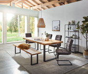 Moderner Esstisch aus Holz mit Metallbeinen, umgeben von gepolsterten Stühlen und einer Bank, in einem hellen Raum mit großen Fenstern und Blick ins Grüne, seitliche Perspektive.