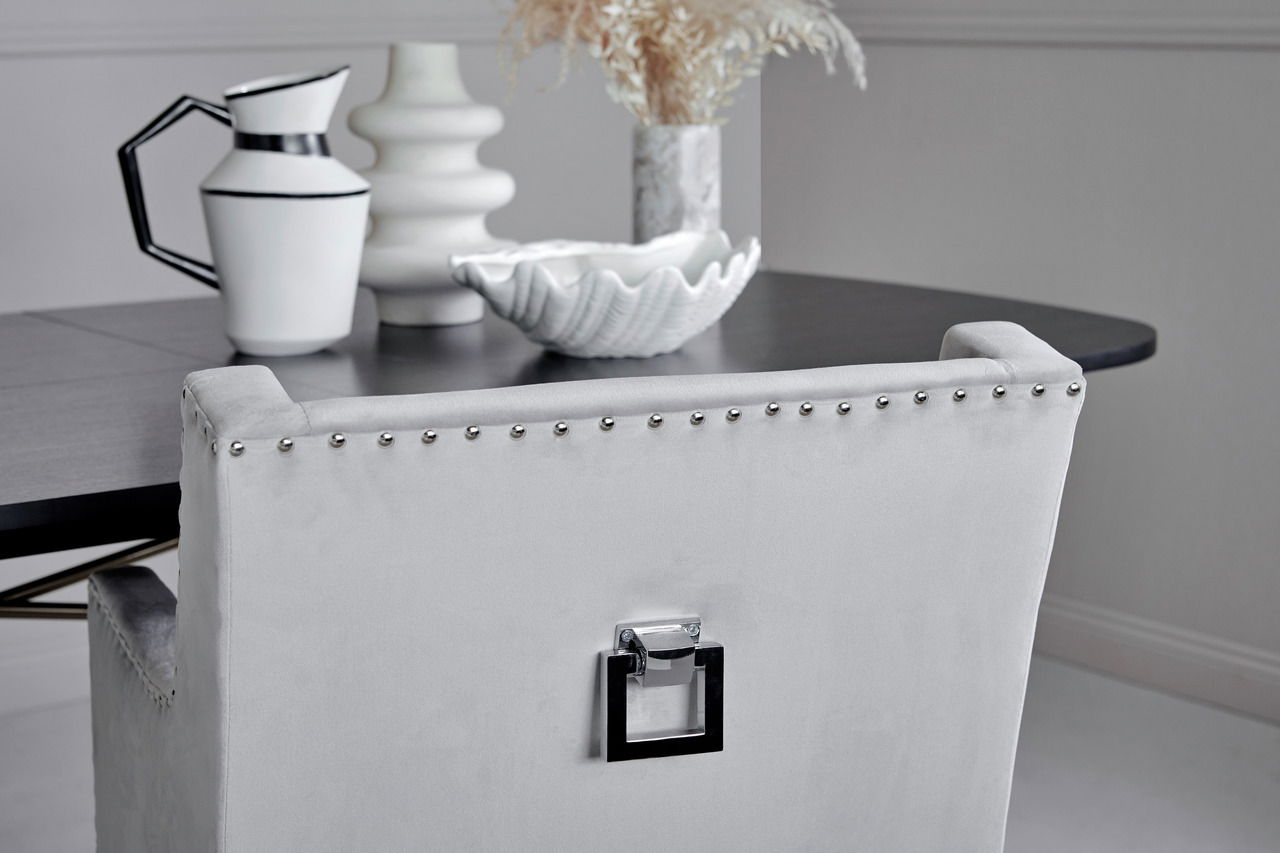 Rückansicht eines eleganten, grauen Stuhls mit Nietenverzierung und quadratischem Griff, vor einem dunklen Tisch mit dekorativen Objekten.