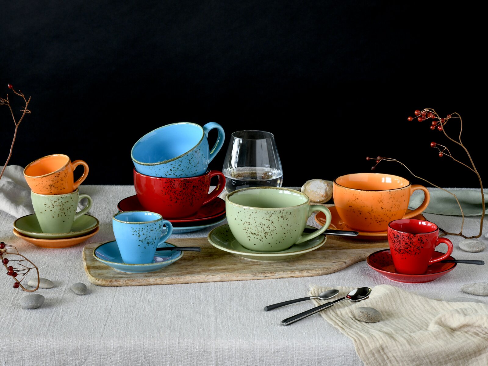 Buntes gesprenkeltes Keramik-Tassen- und Untertassen-Set in Rot, Blau, Grün und Orange auf gedecktem Tisch mit Glas Wasser und Gebäck, fotografiert in leichter Seitenansicht (3/4-Perspektive) vor schwarzem Hintergrund.