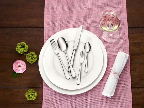 Elegantes Tafelbesteck-Set Oscar, 30-teilig, auf einem gedeckten Tisch mit rosa Tischläufer, Blumen und einem Glas Weißwein, aus der Vogelperspektive.