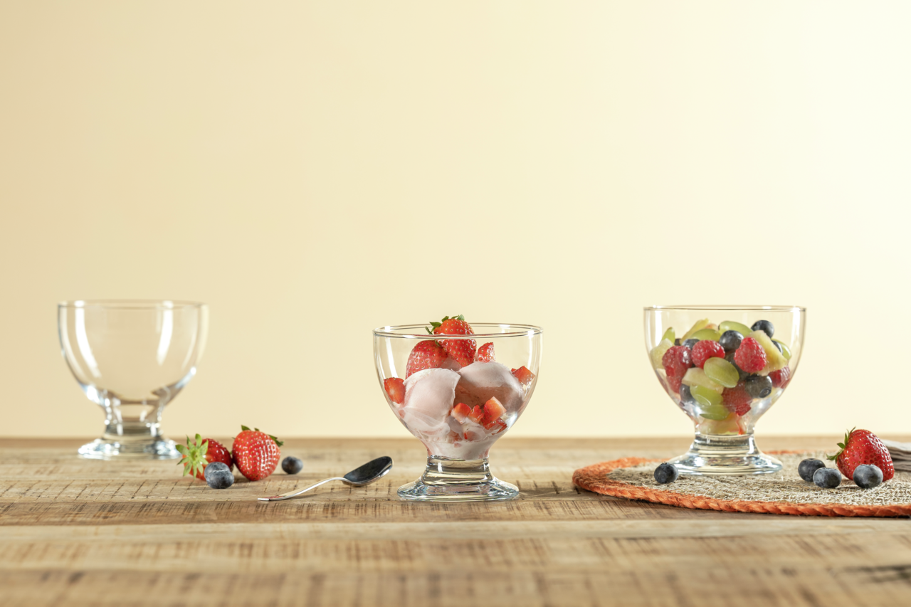 Drei Dessertschalen aus Glas, gefüllt mit frischen Früchten und Eis, auf einem Holztisch. Perspektive von vorne.
