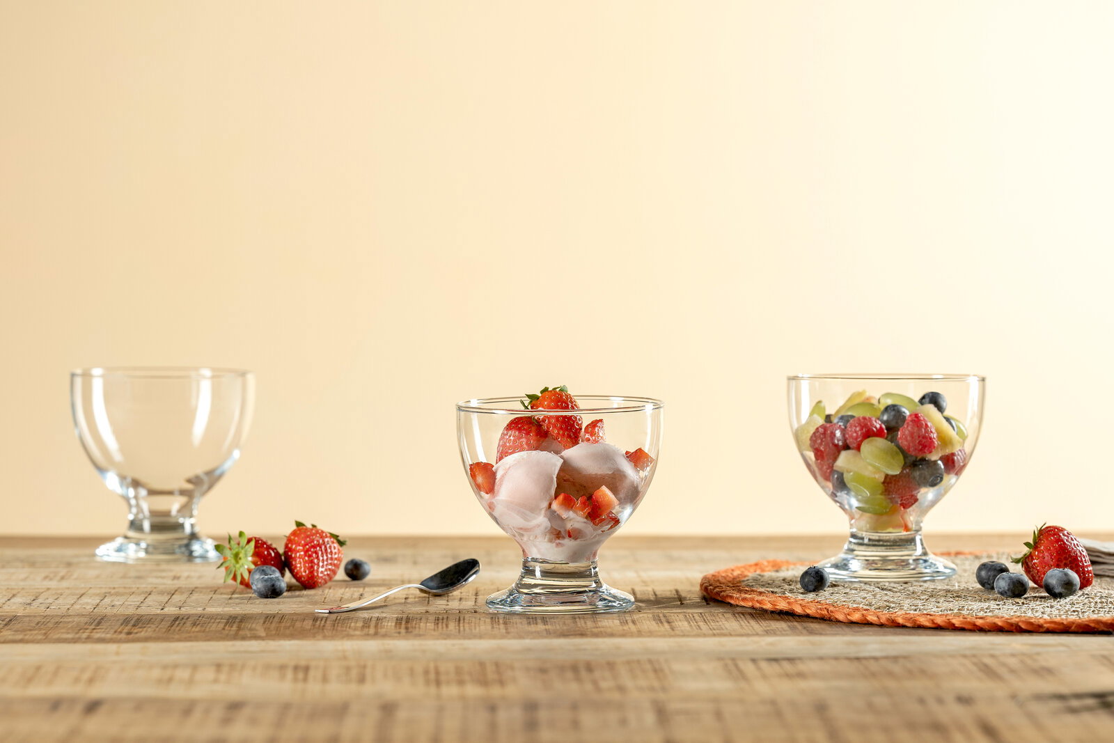 Drei Dessertschalen aus Glas, gefüllt mit frischen Früchten und Eis, auf einem Holztisch. Perspektive von vorne.