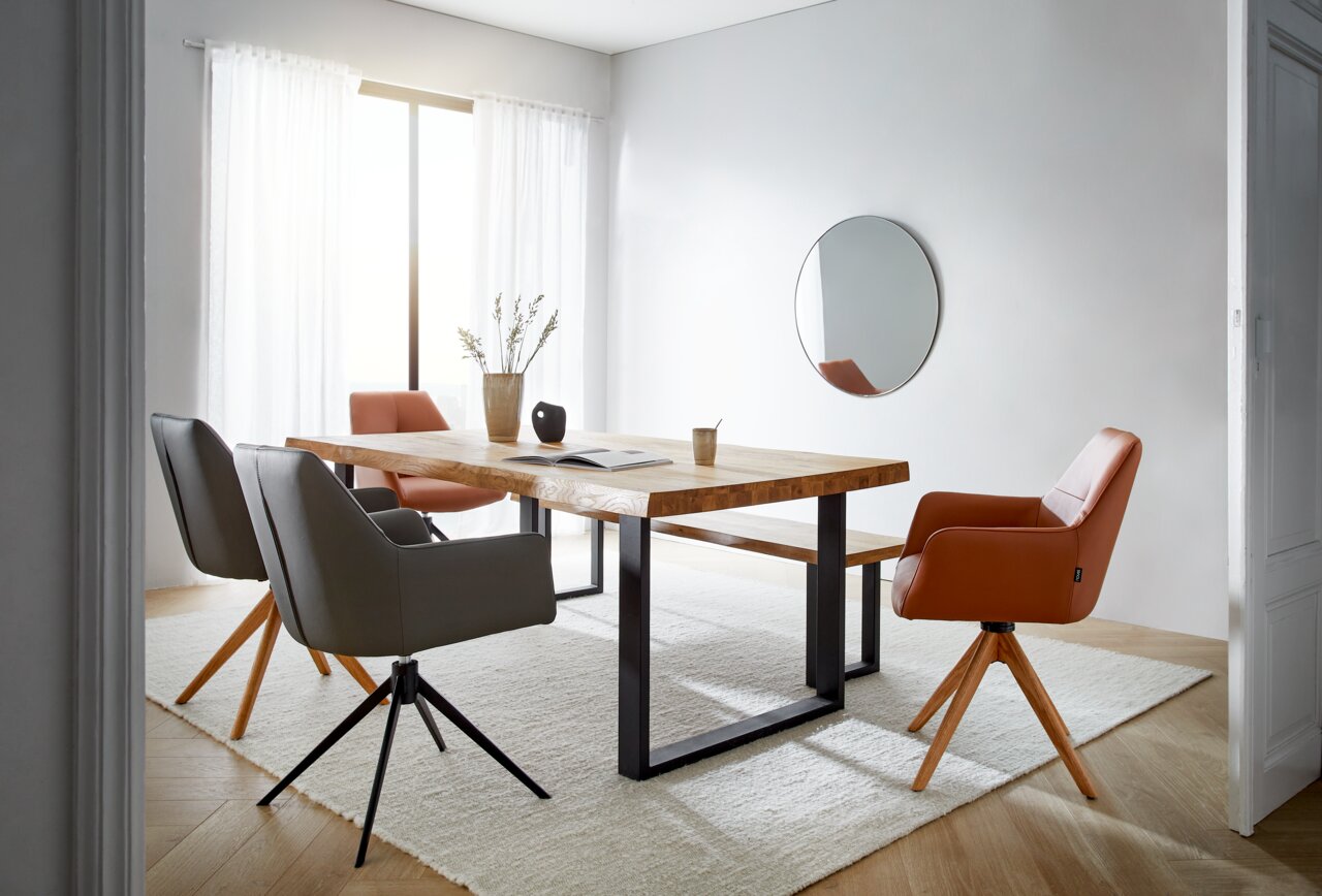 Moderner Esstisch aus Holz mit Metallbeinen, umgeben von stilvollen Stühlen in einem hellen Raum mit großem Fenster, aus seitlicher Perspektive fotografiert.