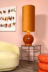 Orangefarbene Stehlampe mit zylindrischem Schirm auf einem runden Glastisch, seitliche Perspektive, im Hintergrund ein pastellfarbener Raum mit einem bunten Vogelbild.