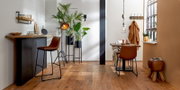 Moderne poef van bruin leer met houten poten, aan de zijkant geplaatst in een stijlvol ingerichte ruimte.