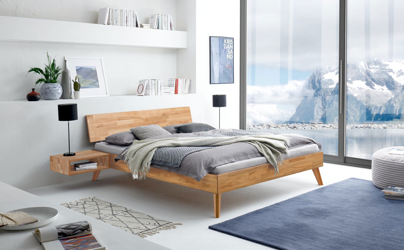 Modernes Bettgestell aus Holz mit integriertem Nachttisch, in einem hellen, stilvollen Schlafzimmer mit Blick auf eine Berglandschaft durch große Fenster. Perspektive von der Seite.