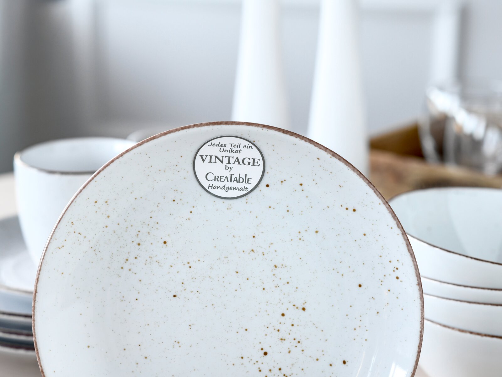 Weißer, gesprenkelter Keramikteller im Vintage-Stil mit braunem Rand und Aufkleber „VINTAGE by CreaTable Handgemalt“, Nahaufnahme von oben/leicht frontal; im Hintergrund unscharf weitere Schalen und Teller.