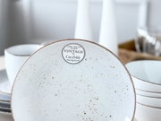 Weißer, gesprenkelter Keramikteller im Vintage-Stil mit braunem Rand und Aufkleber „VINTAGE by CreaTable Handgemalt“, Nahaufnahme von oben/leicht frontal; im Hintergrund unscharf weitere Schalen und Teller.