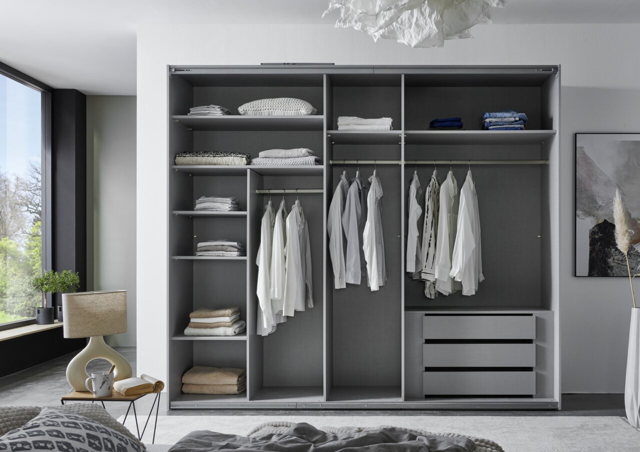 Moderner Schwebetürenschrank mit drei Türen, offene Perspektive, zeigt Innenraum mit Kleiderstangen, Regalen und Schubladen, gefüllt mit Kleidung und Textilien.