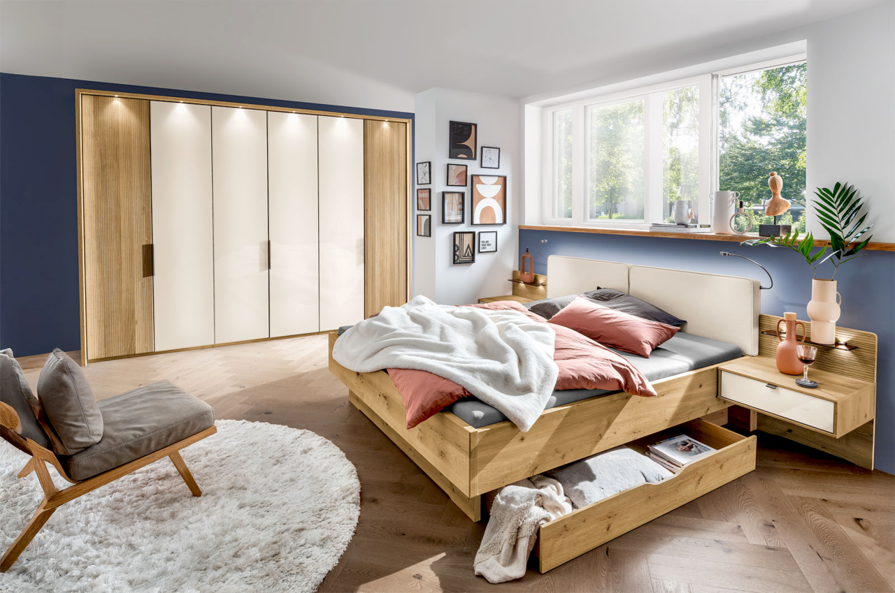 Venda Drehtürenschrank 6-türig NEW YORK Modernes Schlafzimmer mit einem großen, sechstürigen Drehtürenschrank in Holzoptik und weißen Fronten, einem Futonbett mit Bettkasten und Nachttischen, aus der Perspektive von der Zimmerecke aus gesehen.