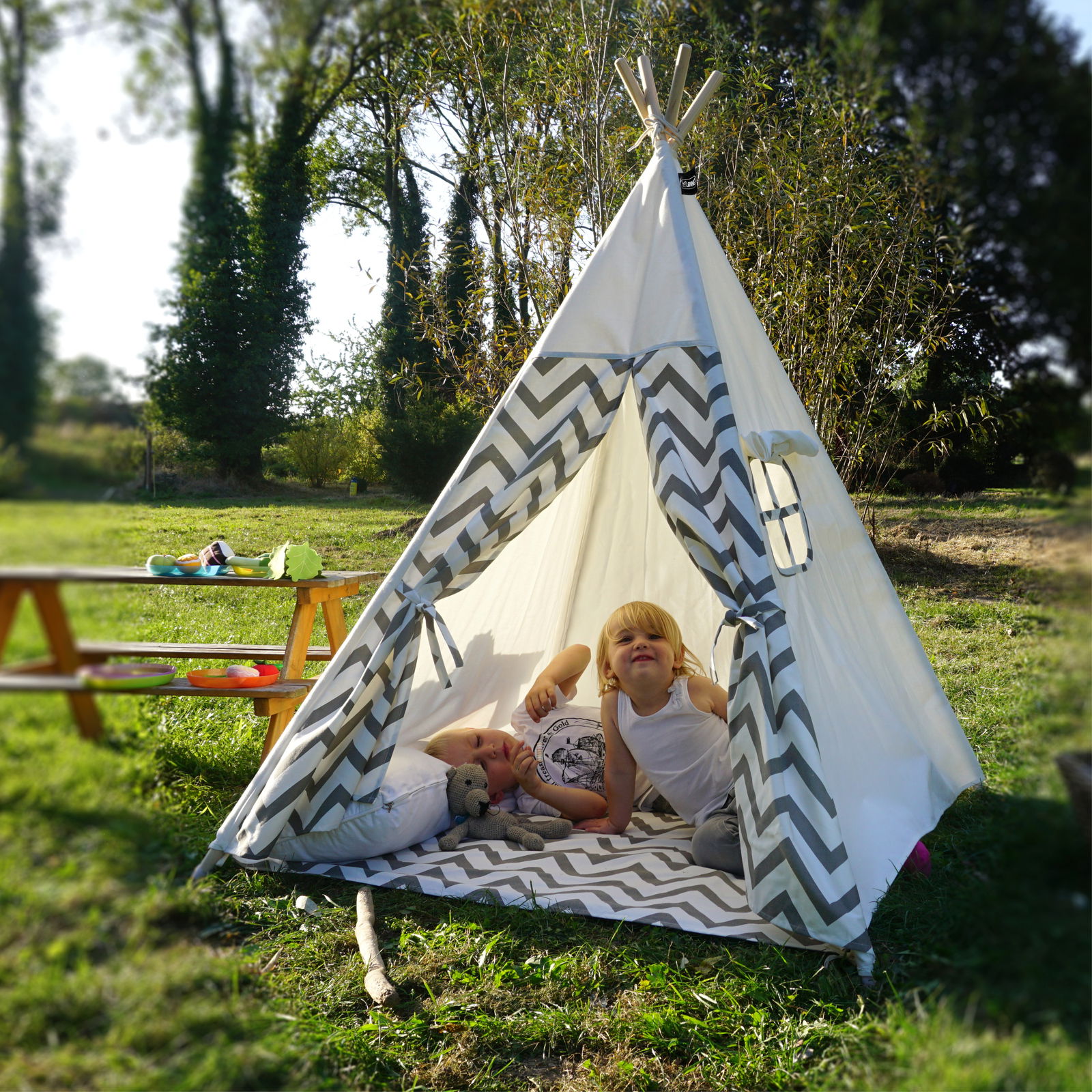 Zwei Kinder spielen in einem grauen Zickzack-Tipi-Zelt auf einer Wiese, frontal fotografiert.