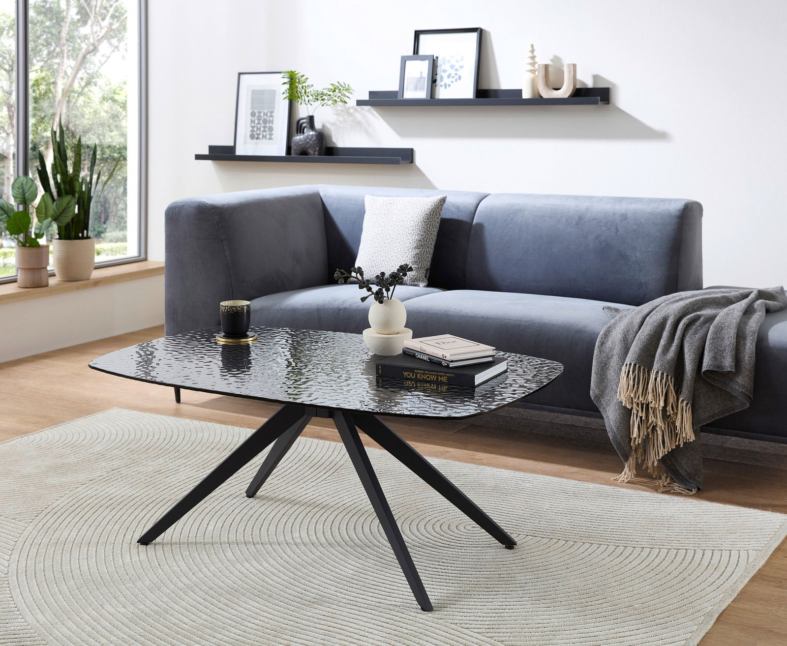 Moderner Couchtisch mit schwarzer Glasplatte und Metallbeinen, im Wohnzimmer mit grauem Sofa und Dekorationen, seitliche Perspektive.