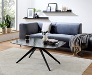 Moderner Couchtisch mit schwarzer Glasplatte und Metallbeinen, im Wohnzimmer mit grauem Sofa und Dekorationen, seitliche Perspektive.