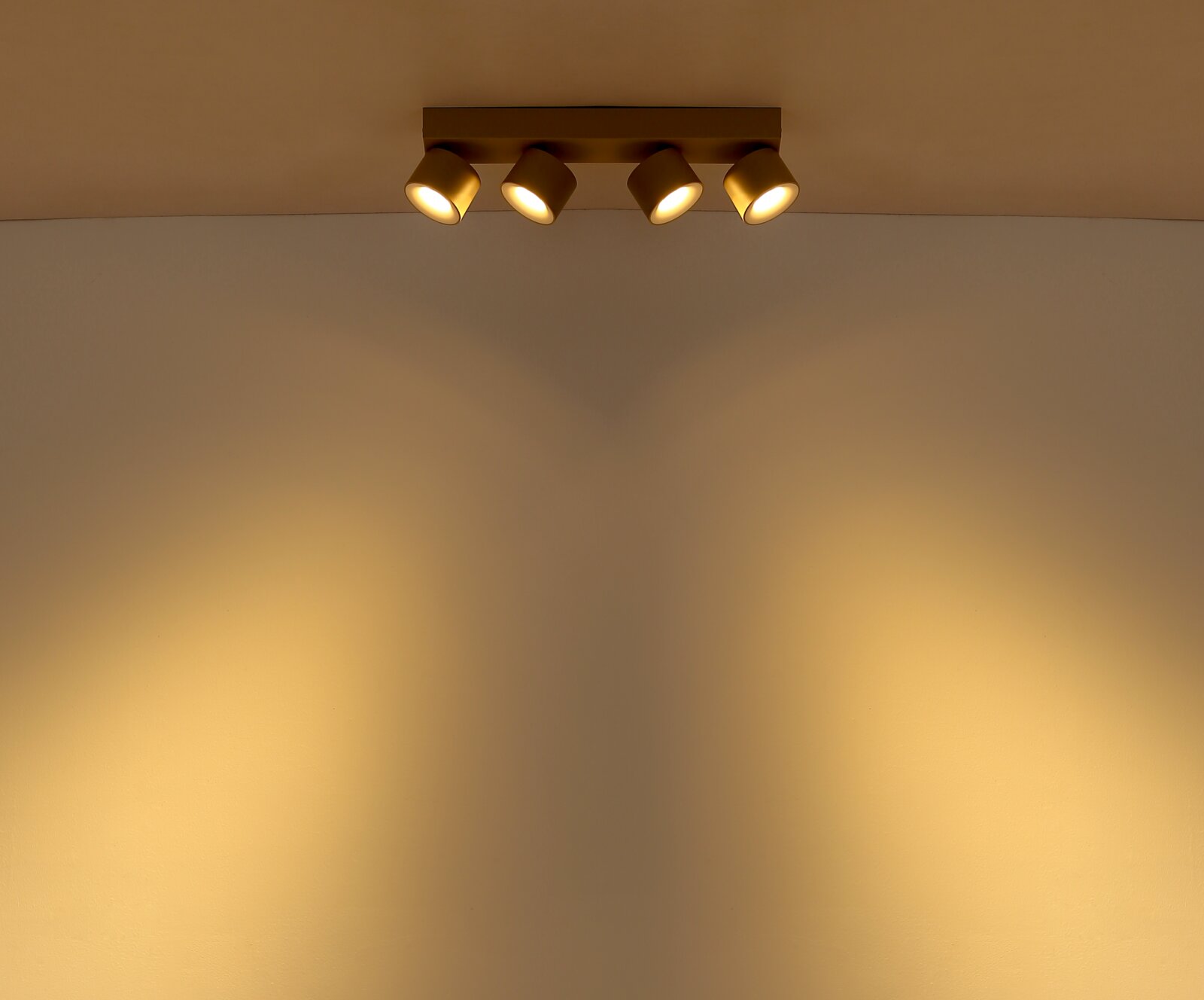 Deckenmontierte 4-flammige Spotleiste in mattem Gold mit warmweißem Lichtkegel an der Wand, Aufnahme aus leichter Untersicht (Blick nach oben)