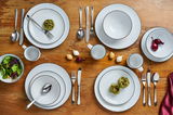 Vogelperspektive auf ein elegantes 24-teiliges Kombiservice 'Black Star' mit schwarzem Rand auf einem Holztisch, inklusive Teller, Schüsseln, Tassen und Besteck, dekoriert mit frischen Zutaten.