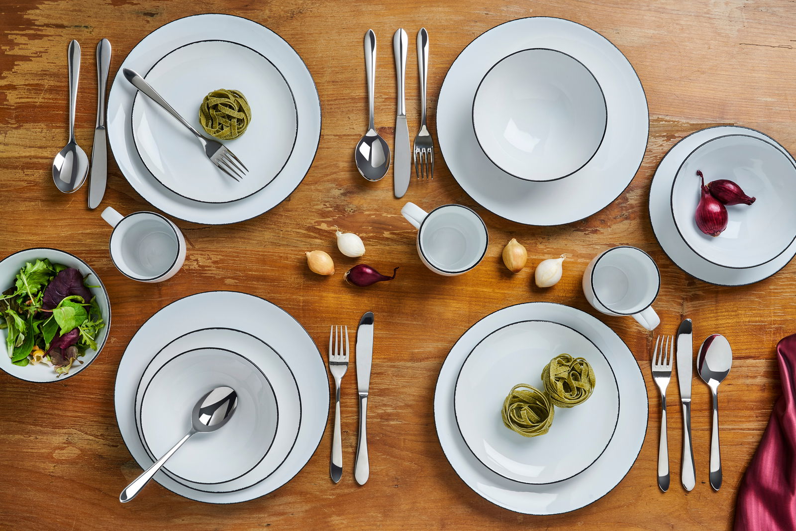 Vogelperspektive auf ein elegantes 24-teiliges Kombiservice 'Black Star' mit schwarzem Rand auf einem Holztisch, inklusive Teller, Schüsseln, Tassen und Besteck, dekoriert mit frischen Zutaten.