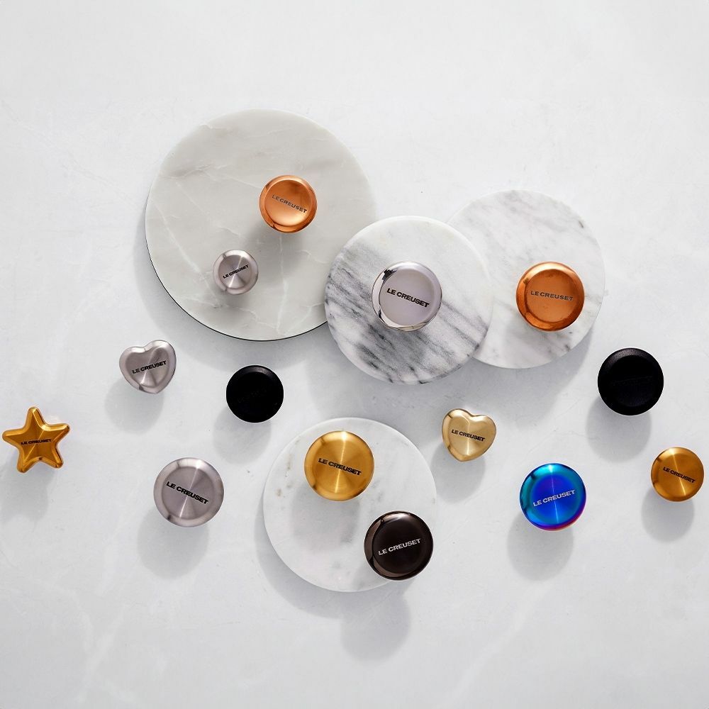 Verschiedene Deckelknäufe aus Edelstahl in verschiedenen Formen und Farben, von oben betrachtet, auf einer weißen Oberfläche arrangiert.